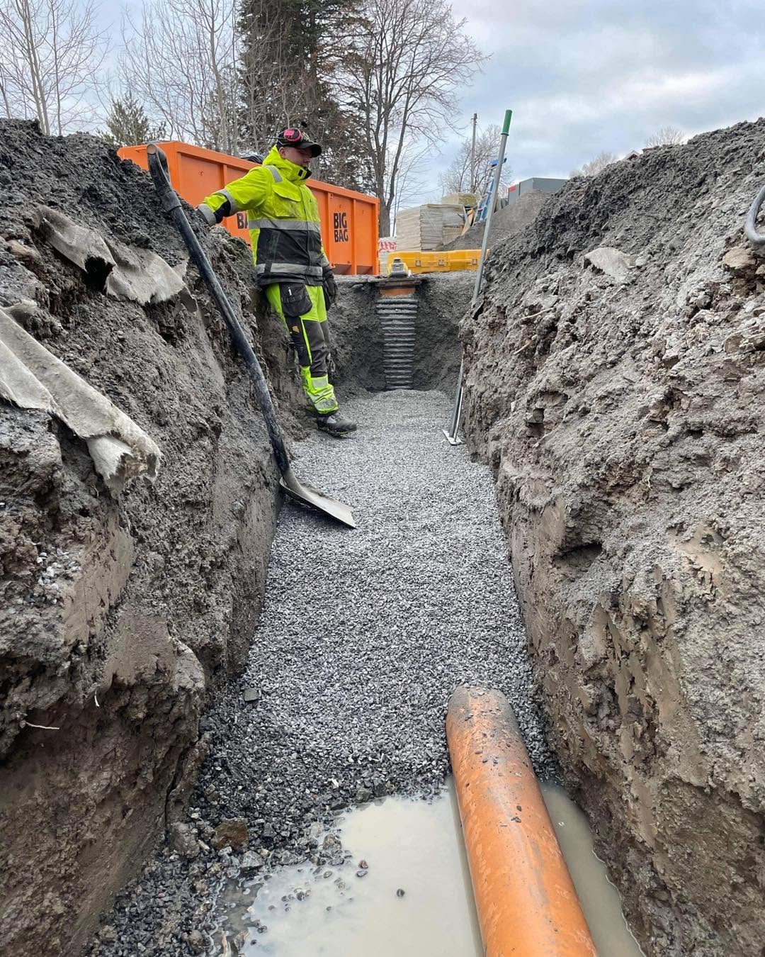 Arbetare i varselkläder stående i ett djupt schakt med grusbädd, orange rör och en schaktbrunn.