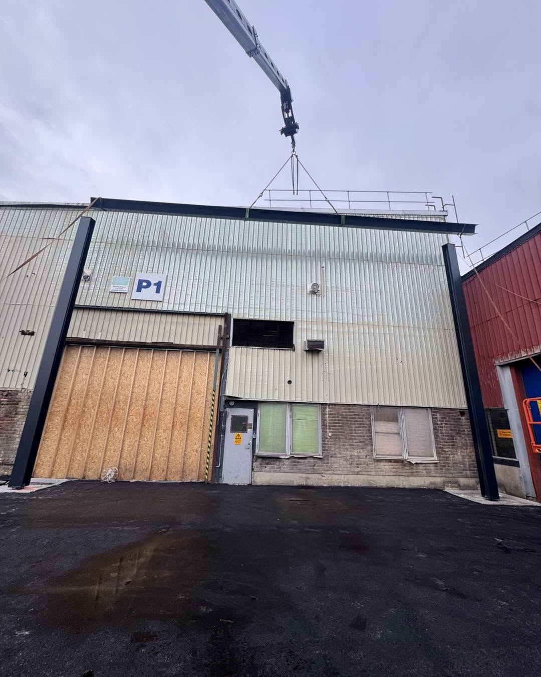 Kran lyfter en lång stålbalk på plats ovanpå två upprestade stålpelare framför en industribyggnad.