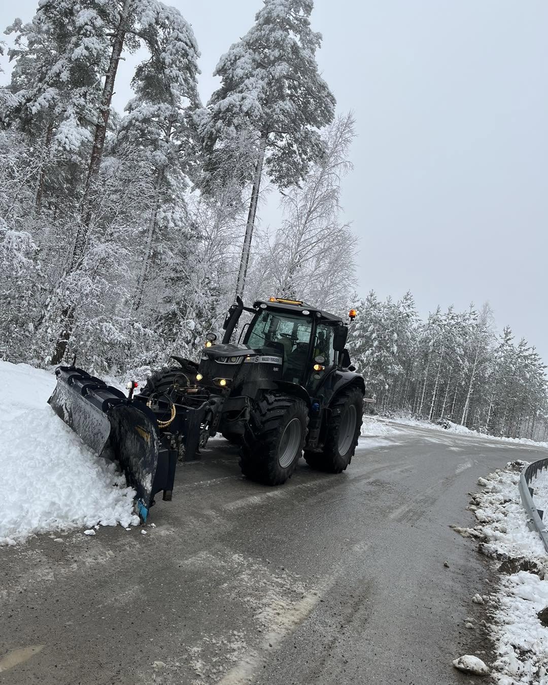 Traktor med snöplog på en väg omgiven av snötäckta träd.