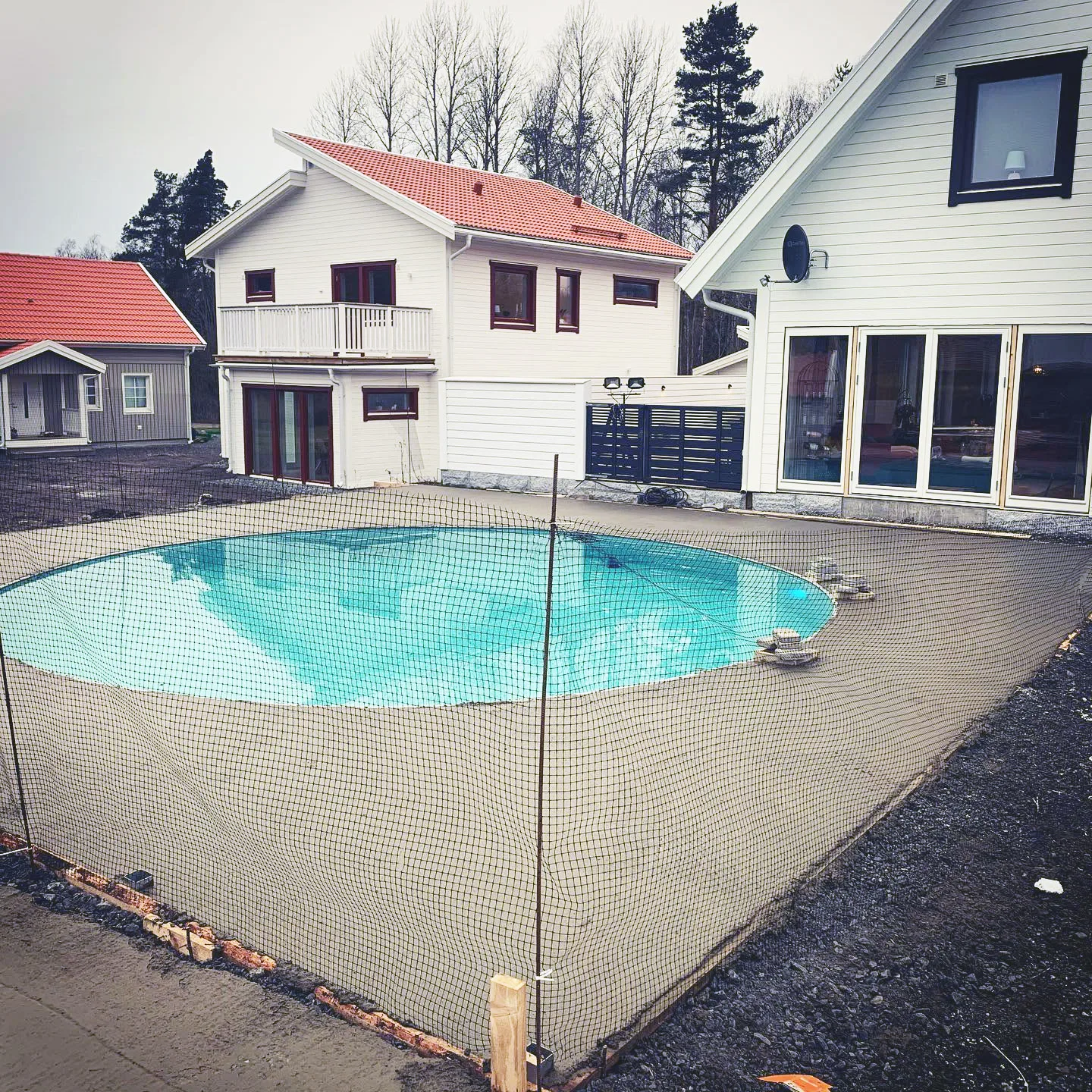 Ingrävd pool med säkerhetsnät och omgivande platsbyggd betongplatta vid bostadshus.