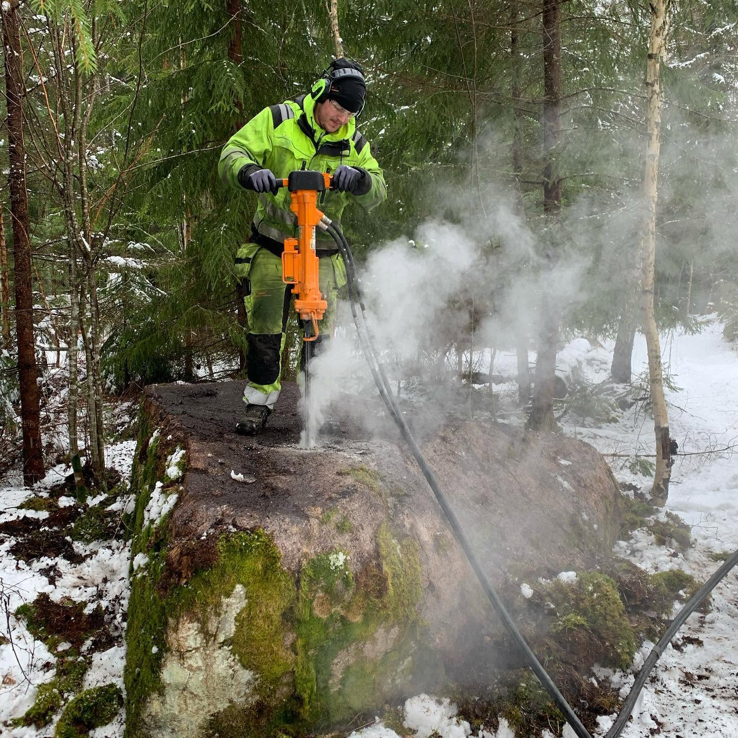 Person i varselkläder, hörselkåpor och skyddsglasögon borrar i ett stort mossigt stenblock med en orange bergborrmaskin. Ett dammoln stiger från borrhålet i skogsmiljö vintertid.