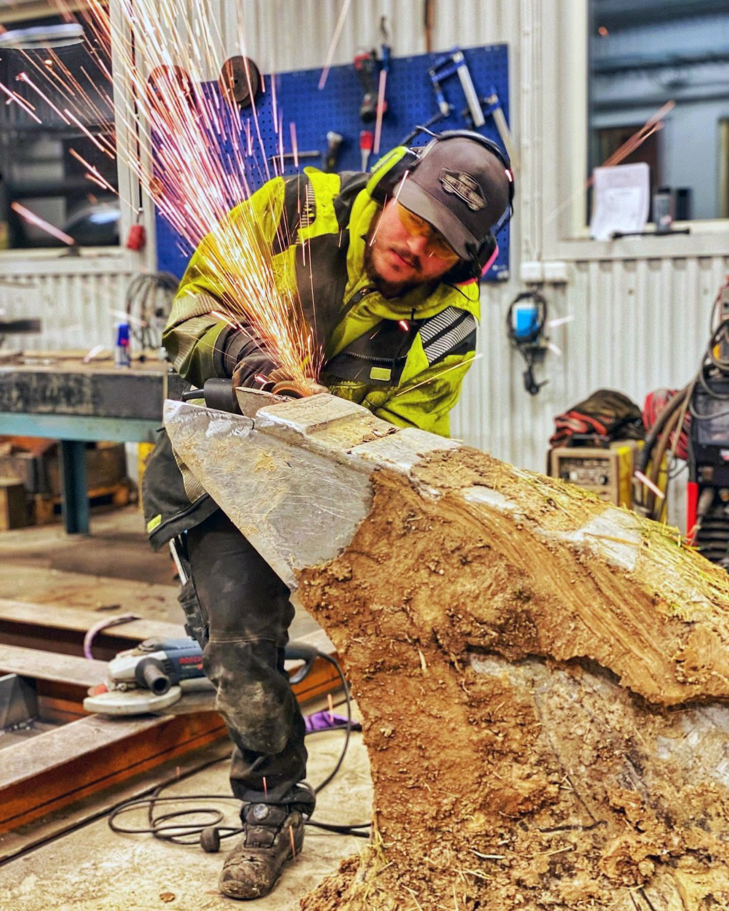 Jocke i varselkläder, skyddsglasögon och hörselkåpor slipar en lerig stålkomponent med vinkelslip. Gnistregn sprutar uppåt i verkstaden.