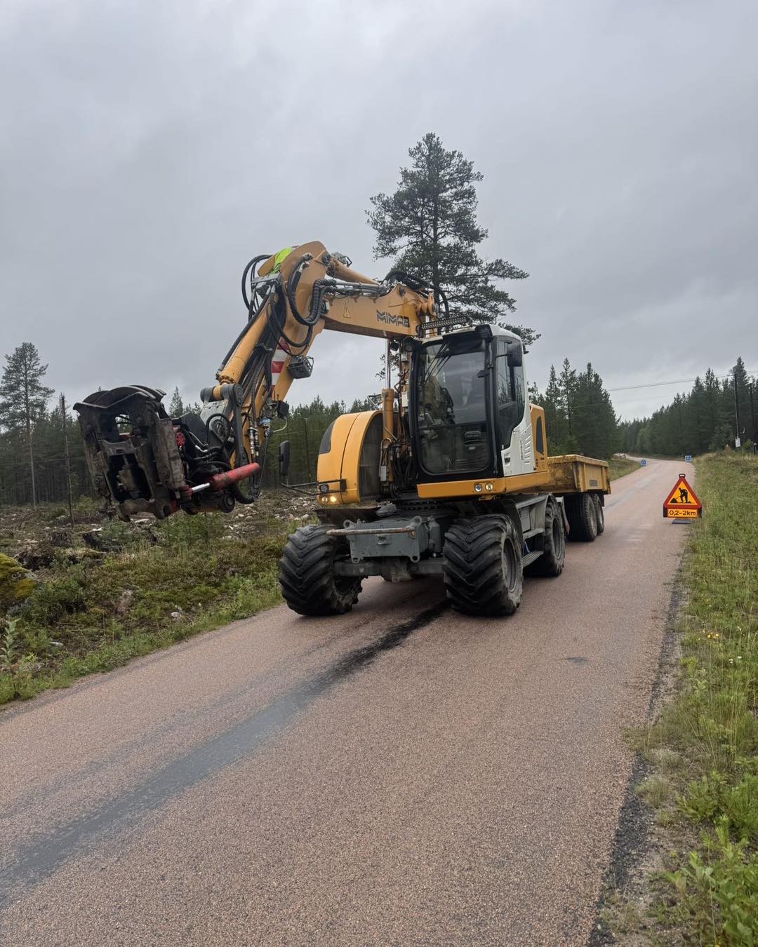 Gul hjulgrävare med släp arbetar vid kanten av en landsväg.