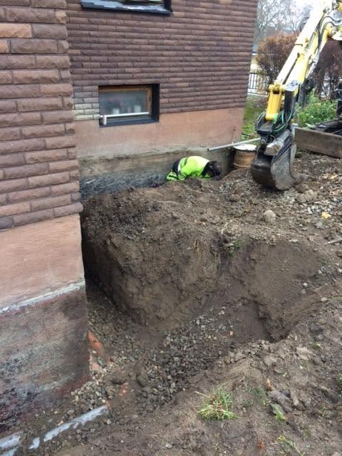 Schaktarbete intill en husgrund med grävmaskin och person i varselkläder.